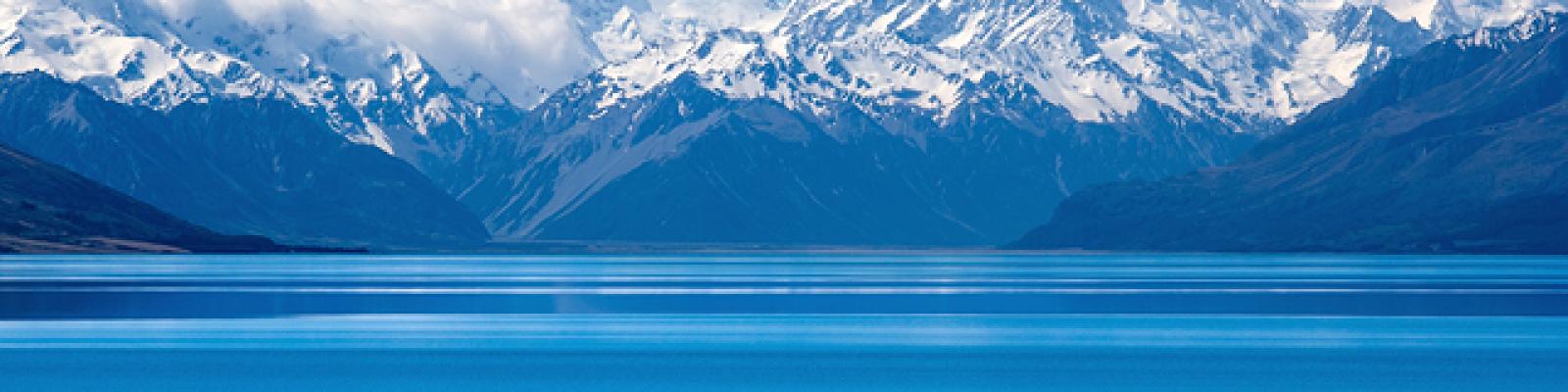 Image of blue bay water in foreground with snow-capped mountains and clouds in the distance